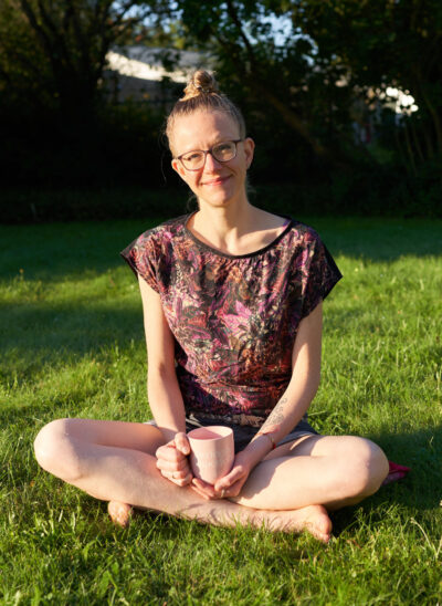 Lisa Jara sitzt mit gekreuzten Beinen auf einem grünen Rasen und lächelt mit leicht geneigtem Kopf und freundlichem Gesicht in die Kamera. In den Händen hält sie einen rosa-farbenen Becher mit der Aufschrift "Perfectly Imperfect" (Englisch für "perfekt unperfekt")
