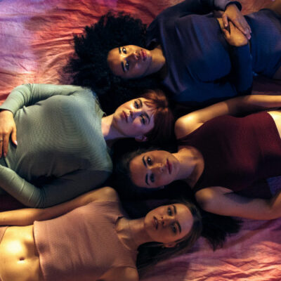 Bild mit vier jungen weiblich gelesenen Personen mit verschiedenen Hautfarben, Haarfarben und Kleidung, die auf dem Rücken liegen, die Köpfen aneinander und selbstbestimmt in die Kamera schauen
