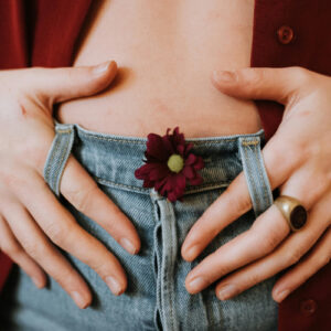 Photo of the lower abdomen of a white person in blue jeans, wearing a red, open blouse and a dark red flower in the jeans' button hole, hands at their side, forefingers casually woven through the belt loops