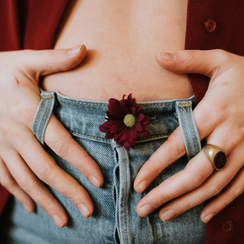 Foto des Schoßraums einer weißen Person in Blue Jeans mit roter, offener Bluse und einer dunkelroten Blume im Knopfloch der Jeans, die Hände locker an den Seiten, die Zeigefinger lässig in den Gürtelschlaufen eingehängt