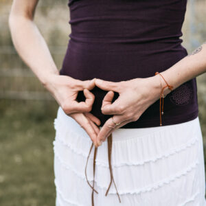 Consultation-Tile Image of the hands and arms of a white person dressed in a dark purple shirt and white skirt holding their hands in a downward-facing triangle in front of their womb space