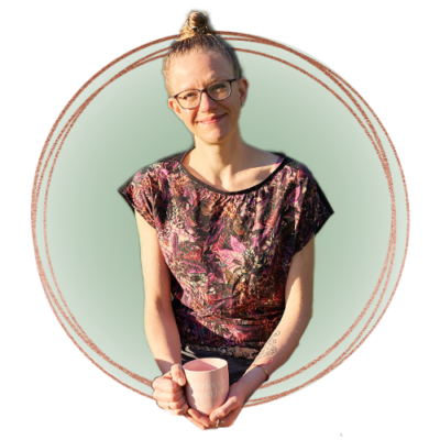 Lisa Jara smiles into the camera with a lightly tilted head, holding a soft pink coffee mug saying "Perfectly Imperfect!". Behind her is a light green background, and a frame of three rose-gold coloured circles surrounds her.