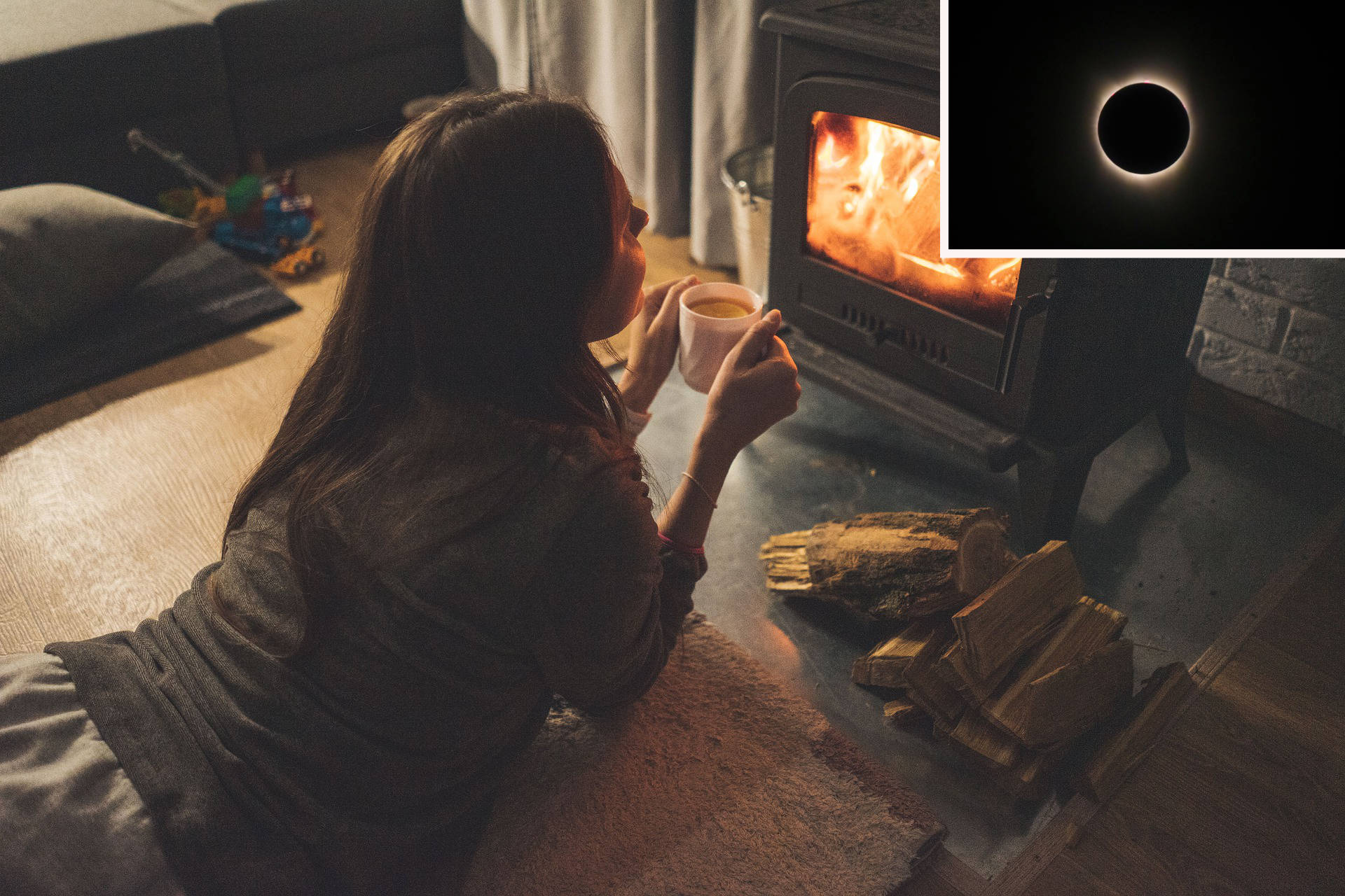 Photo of a person with long dark-brown hair in a grey shirt and grey pants, lying on their belly in front of a stove; propped up on their elbows, they hold a white cup in their hands, watching the fire; logs are stacked to the right of the stove, in the left background there's a dark green couch; in the upper right corner, there's an image of a dark moon with a corona of light