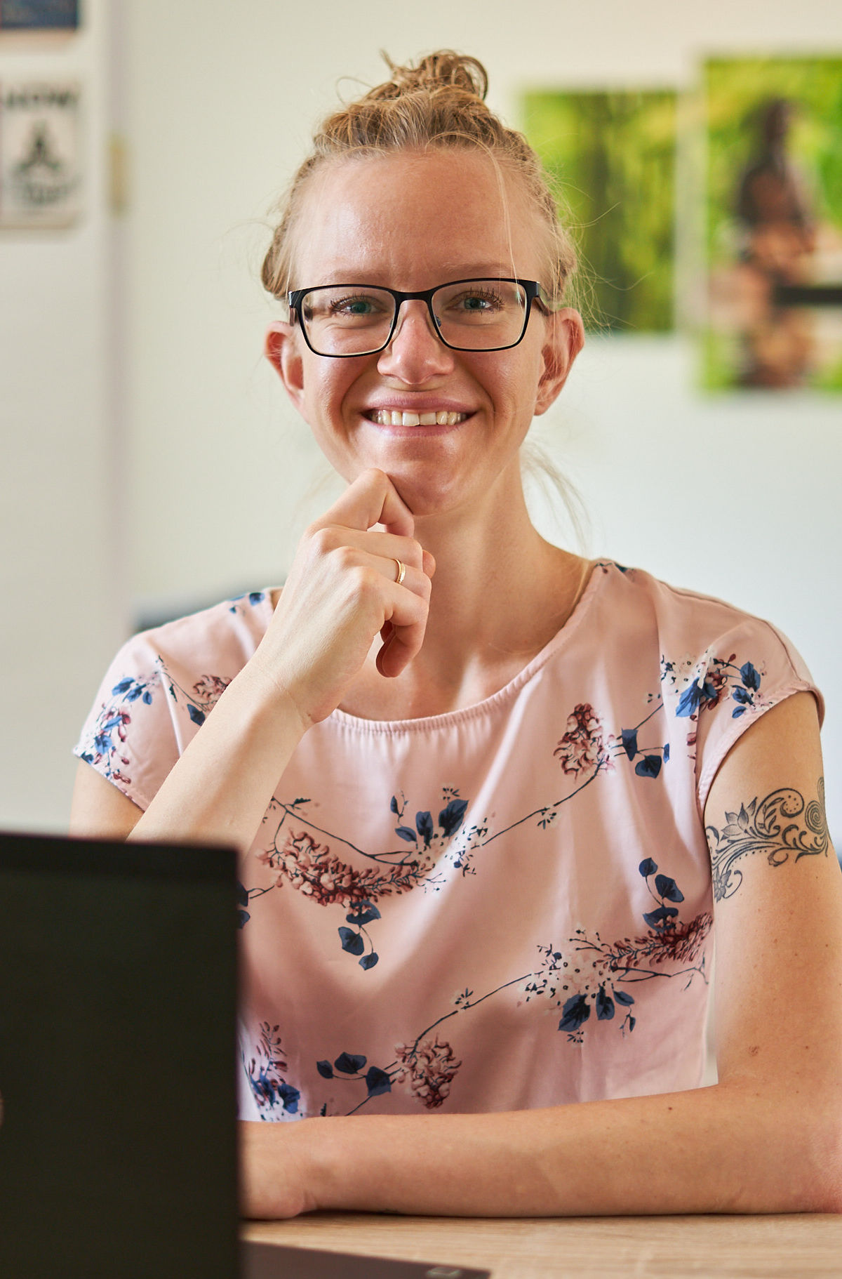 Portrait von Lisa Jara, einer weißen, blonden Frau, mit auf dem Kopf zu einem Dutt zusammengerollten Haaren, in einem rosafarbenen Shirt mit floralem Muster an einem Schreibtisch sitzend, schwarzes Notebook seitlich vor ihr, Kinn leicht auf der rechten Hand aufgestützt; dabei lächelt sie fröhlich in die Kamera