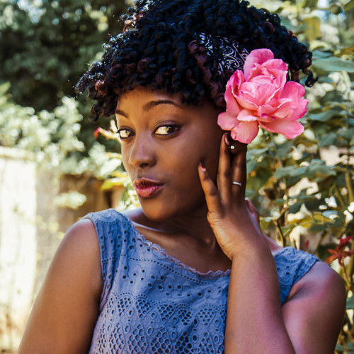 Tile Life Transitions “Colourful Woman” Photo of a black person with curly black hair tied in a ribbon, wearing a blue shirt and a bright pink flower in their hair, left hand on their left cheek, looking into the camera with the hint of a smile