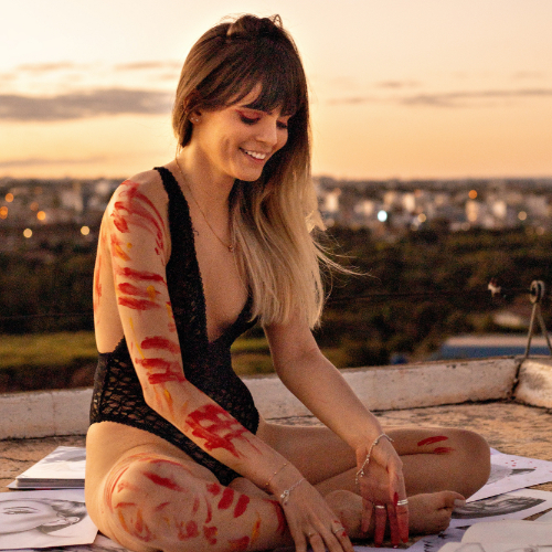 Tile Menstrual Health “Woman with red symbols” Photo of a white woman with long hair lying loosely over her left shoulder; she's sitting cross-legged on a rooftop, dressed in a black laced bodysuit, red symbols painted all over her arms and legs; she's smiling down at a bunch of black-and-white drawings strewn around her