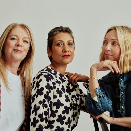 Tile Perimenopausal Health “3 Women” Photo of three people perceived as women in midlife; to the left a white person with long, blond hair, dressed in a white shirt and smiling slightly into the camera; in the middle a light brown person with short white-grey hair, dressed in a black-and-white-patterned shirt, looking confidently into the camera; to the right a white person with medium length hair, dressed in a denim shirt, chin propped up on their right hand, looking over to the other two people