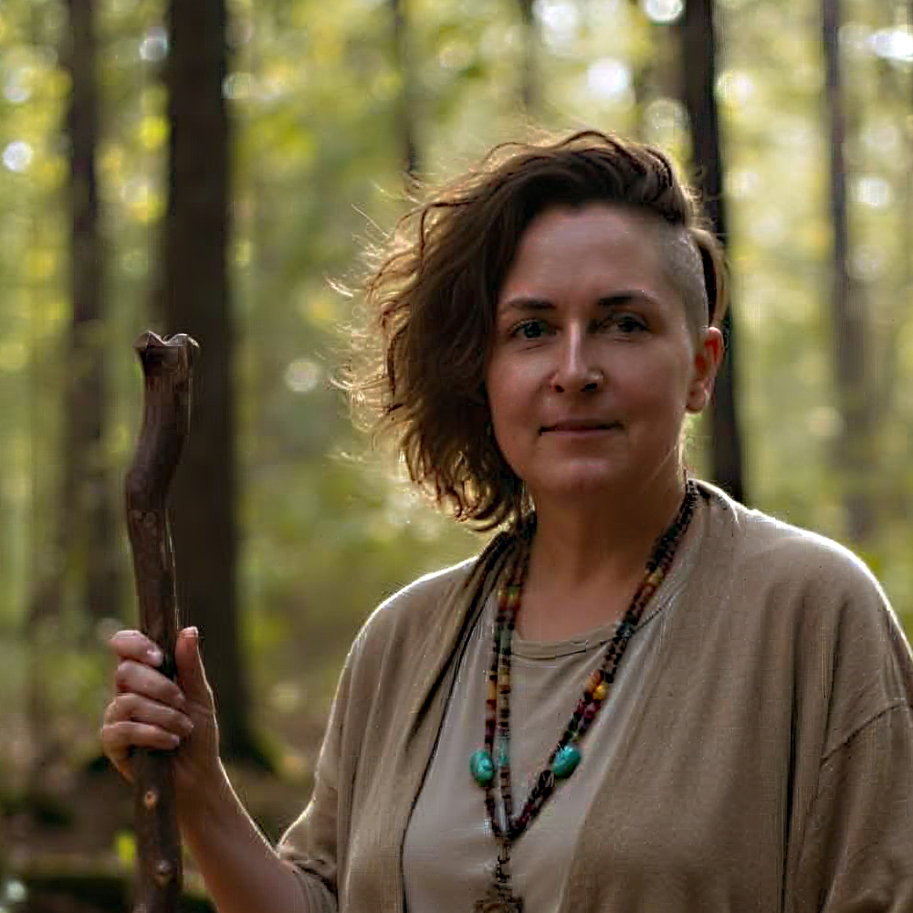 Ixchel Lunar with staff Photo of Ixchel Lunar (they/them), a white-presenting, indigenous person with brown hair combed to their right and an undercut on the left, wearing beige-brown loose clothes and turquoise jewellery, holding a wooden staff in their right hand, looking straight into the camera