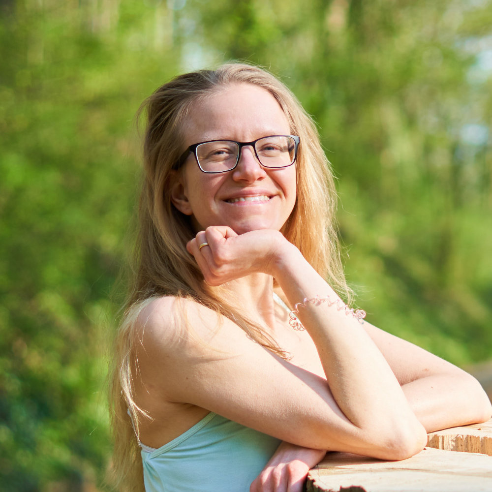 Lisa Jara close-up green Photo of Lisa Jara (she/her), a white person with long blonde hair falling loosely over her shoulder, wearing a turquoise top, right arm propped up on a tree stump she's leaning against, smiling brightlx into the camera