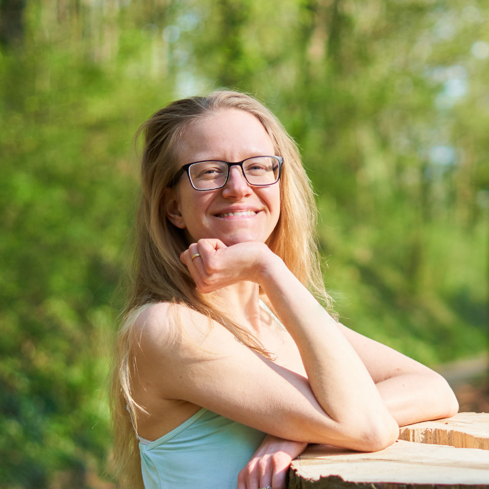 Photo of the head and upper torso of Lisa Jara, a blond, white woman with glasses in a turquoise tank top, head propped up on her right arm laying on the tall stump of a tree, hair falling openly over her shoulders, smiling brightly into the camera; green shrubs in the background