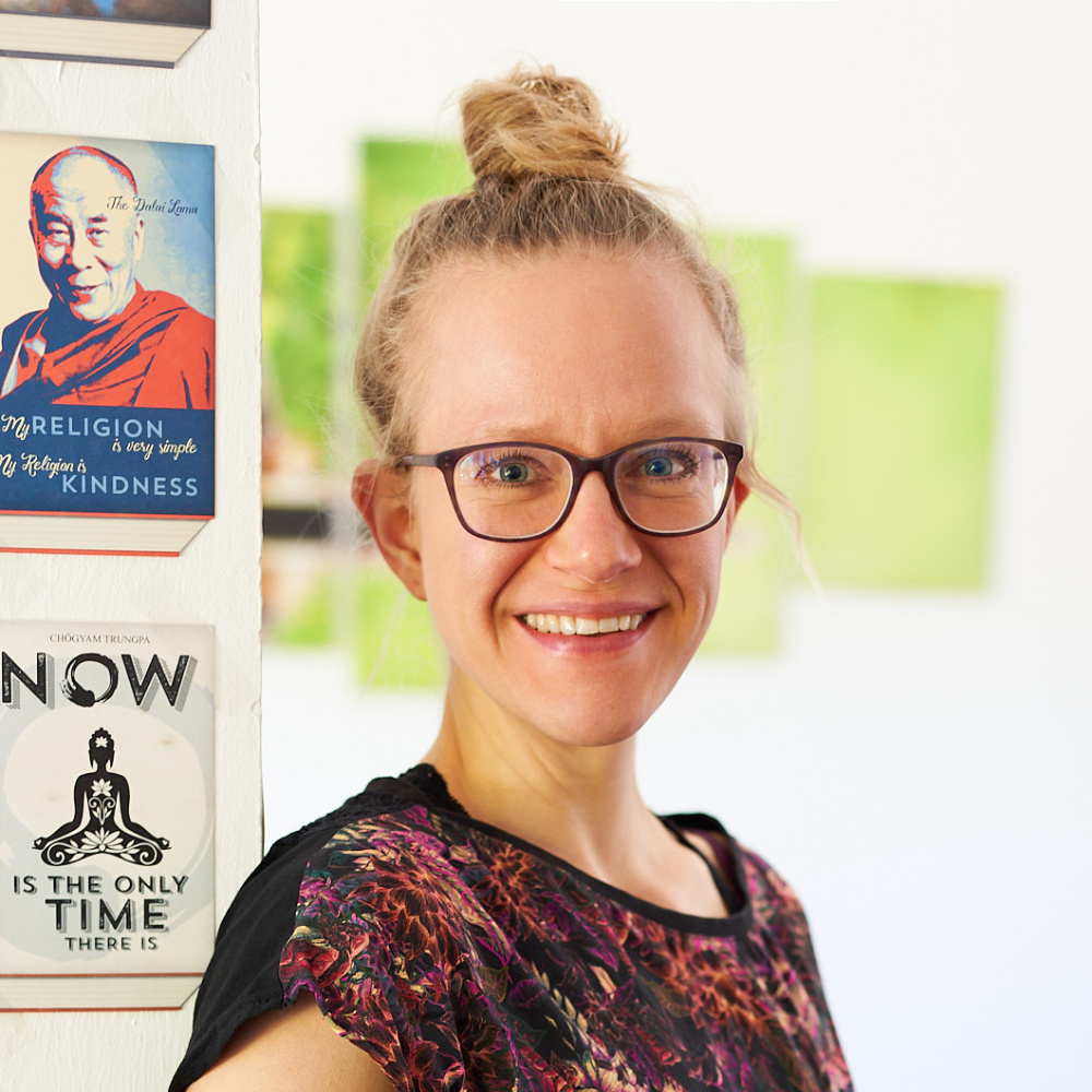 Close-up photo of Lisa Jara, a blond, white woman with glasses, hair in a bun at the top of her head, wearing a dark pinkish shirt and smiling brightly into the camera; the background shows a green wall art and two postcards, one with the saying "Now is the only time there is." and "My religion is kindness."
