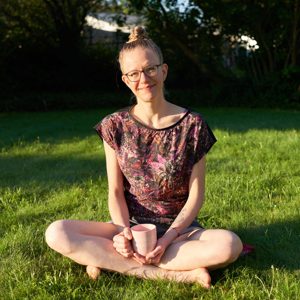 Photo of Lisa Jara, a blond, white woman dressed in a dark pink-ish shirt and grey shorts, sitting cross-legged on a green lawn, smiling into the camera with a lightly tilted head; in her lap, she holds a soft pink coffee mug saying "Perfectly Imperfect!"