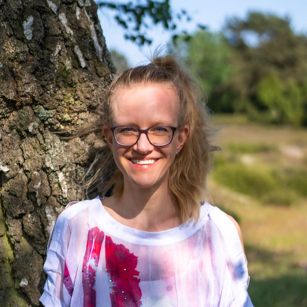 Close-up photo of Lisa Jara, a white, blond woman with glasses looking and smiling straight into the camera; she's dressed in a white shirt with colourful splashes, hair in a high pony tail falling over her left shoulder, back leaning against a tree trunk
