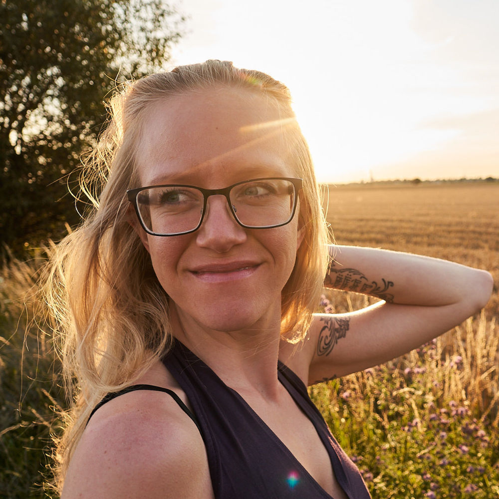 Close-up photo of Lisa Jara, a white, blond woman with glasses, face turned towards the camera, but gazing to one side, slightly smiling to herself, holding her hair up with her left hand; the setting sun in the background, casting a soft golden light over the scenery