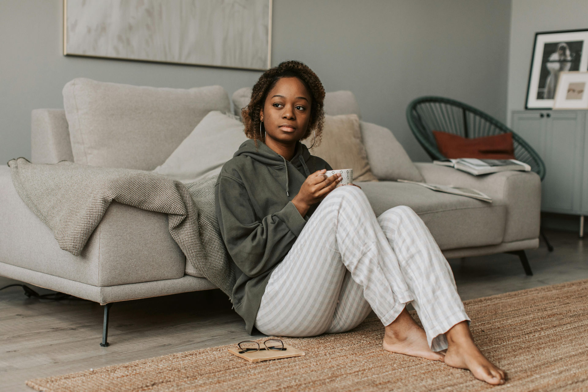 Foto einer Person of Colour gekleidet in ein weites, olivgrünes Sweatshirt und eine weiß-graue lange Pyjama-Hose, die mit leicht angezogenen Knien vor einem Sofa in einem ordentlich aufgeräumten Wohnzimmer sitzt, mit dem Rücken gegen das Sofa gelehnt, Becher in der Hand, nachdenklich in die Ferne blickend